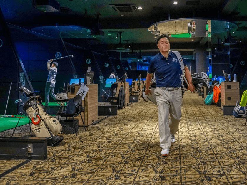 A man in a golf simulation facility walks while carrying clubs; another person swings at a simulator in the background.